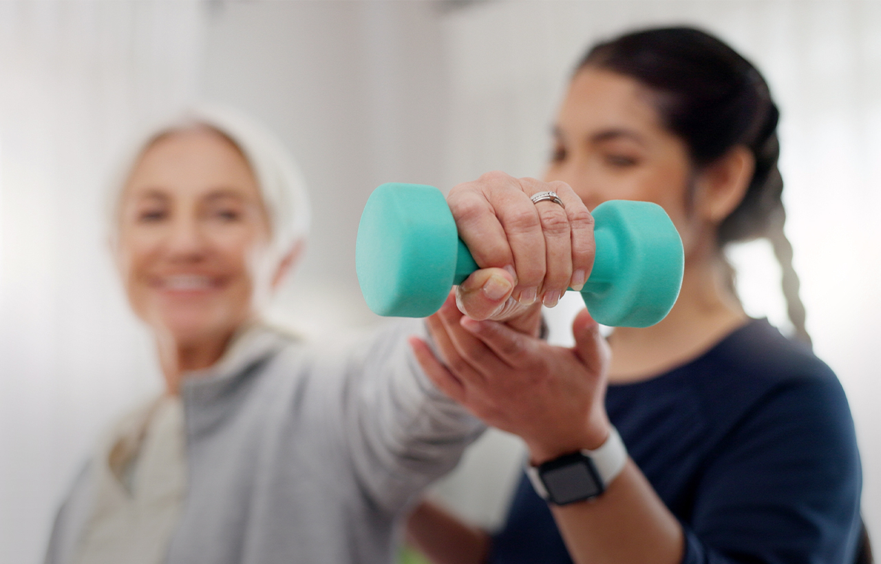 Ältere Dame hält ein Gewicht in der Hand und eine Physiotherapeutin unterstützt sie dabei